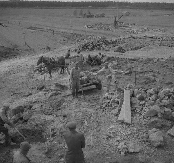 Urjalan Kunnalliskodin rautakautisia röykkiöitä tutkitaan vuonna 1953. Kunnalliskodin rakennustöiden yhteydessä maata oli poistettu räjähdyspanoksin, jolloin esiin oli lennähtänyt kaksi 600-700-luvulle ajoitettavaa keihäänkärkeä, väkipuukko, paimensauvaneula ja kilvenkupura. Jorma Leppäaho saapui paikalle ja kaivoi röykkiön, joka kuuluu samaan laajaan kalmistoalueeseen Ali-Uotilan röykkiöiden kanssa. Leppäaho julkaisi tutkimuksista artikkelin vuonna 1954 Lounais-Hämeen kotiseutu- ja museoyhdistyksen vuosikirjassa. Kuva: Pekka Kosonen(?).