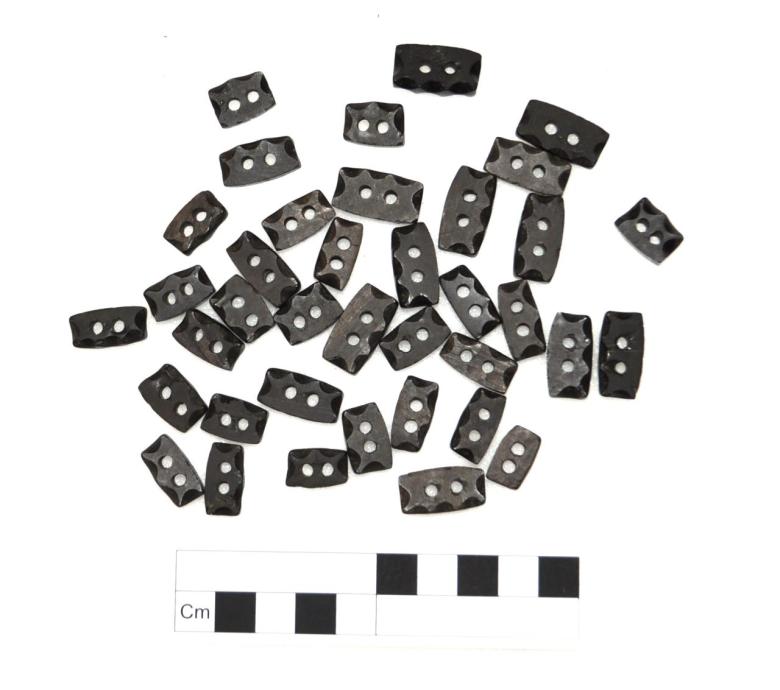 Thirty-eight individual jet beads making up a single composite bracelet. Each bead is a curved-rectangular frame, twice perforated on the long side, and faceted at the edges. Located on the left arm of with skeleton 395.