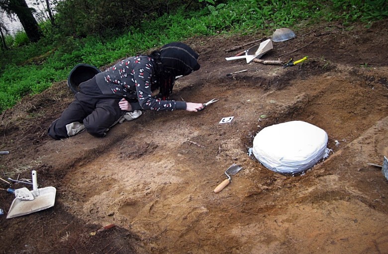 Artikkelin kirjoittaja kaivauksilla. Vieressä tuoreeltaan kipsattu ”Pää”. Kuva: arkeologian laboratorio/Oulun yliopisto.